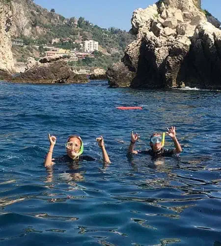 Immagine di due persone che fanno snorkeling in acque cristalline vicino a scogli e coste rocciose.