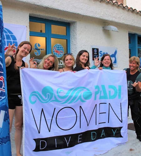 Gruppo di donne sorridenti con bandiera PADI Women Dive Day davanti a una sede di immersioni.