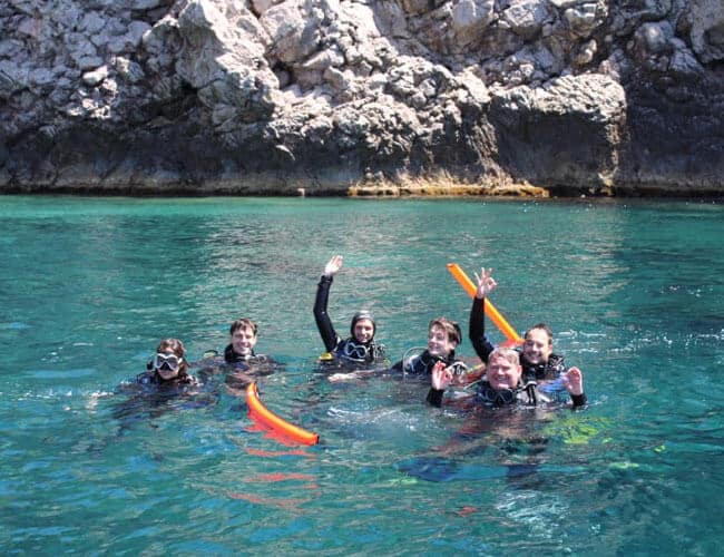Subacquei in acqua con attrezzatura da immersione, sorridenti e felici, vicino a scogliera rocciosa.